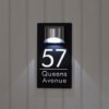 Black LED house number plaque 57 Queens Avenue, lit custom sign on light grey wooden wall